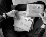 Ręce trzymające stos brązowych kopert, każda z adresem i znaczkiem pocztowym. Wygląda jak przygotowanie do wysyłki listów poleconych ekonomicznych.