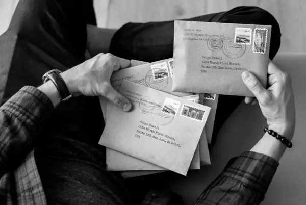 Ręce trzymające stos brązowych kopert, każda z adresem i znaczkiem pocztowym. Wygląda jak przygotowanie do wysyłki listów poleconych ekonomicznych.